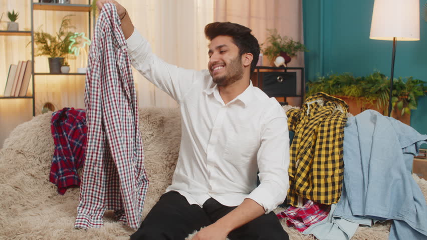 Indian young man at home looking around in confusion, searching through a messy pile of clothes for a shirt. Arabian guy sitting on sofa, feeling frustrated while trying to find something to wear