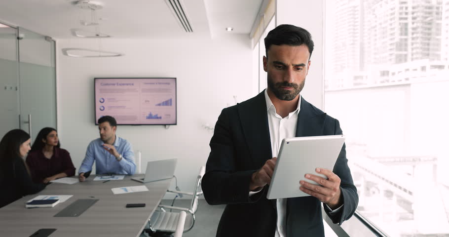 Focused businessman in formal suit standing in conference room using digital tablet, reviewing online reports, analyzing received data, or managing tasks. Innovative tech, workflow using modern device