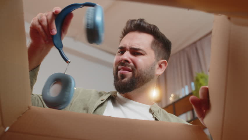 Dissatisfied annoyed male customer open cardboard box receive damaged broken wireless headphones, shocked young man consumer having problem complaint frustrated with bad order delivery at home room.
