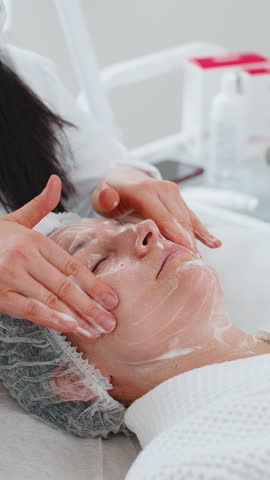 Beautician applying moisturizing medical chemical peeling cream scrub mask on young adult woman face with hands. Girl skincare spa procedure. Facial cosmetology treatment skin regeneration. Vertical