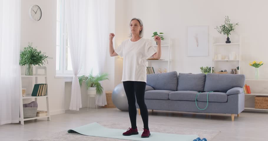 Active sporty healthy senior woman in comfortable clothes standing on an exercise mat in the living room and doing elbow to knee crunches. Sport, fitness, home workout concepts