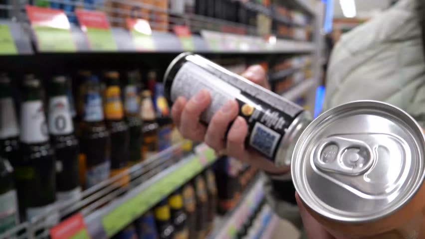 Close-up of female hands holding two cans of alcohol against the background of a shelf with alcohol in a store. Young woman reads the description on cans of alcoholic and non-alcoholic beer, reads the