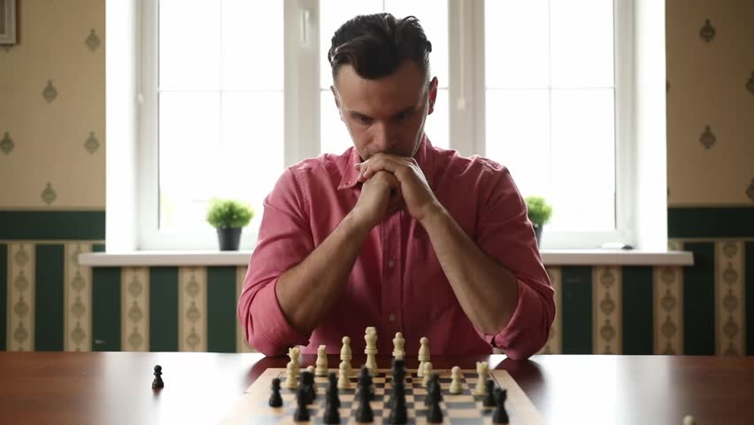 A man in a pink shirt is sitting at a wooden table, deeply focused on a game of chess, with his hands clasped in front of him, contemplating his next move. The background features a bright window.