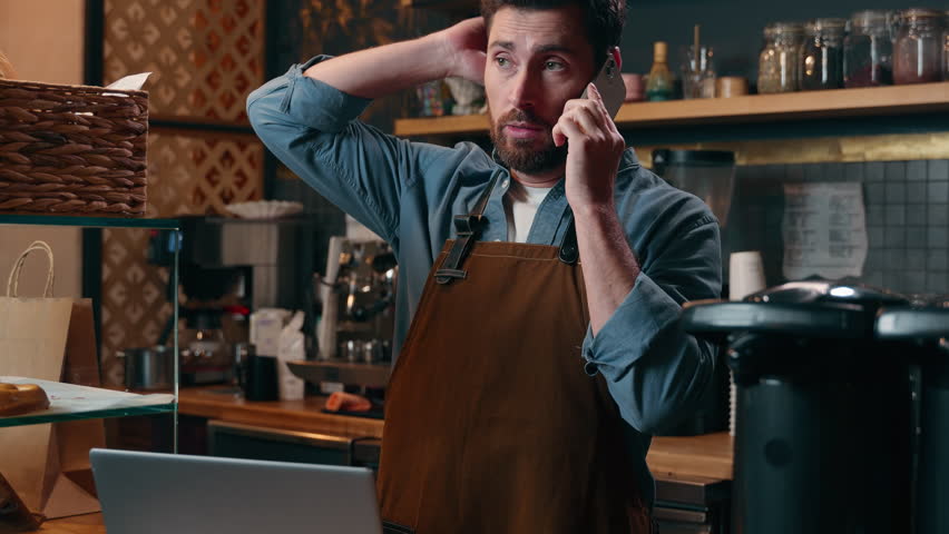 Tired Caucasian guy worker waiter man talk with client mobile phone executive discuss barista seller delivering in cafeteria male typing laptop small business owner placing order online cafe industry