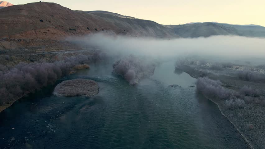 A foggy mountain range with a river running through it. The water is calm and the sky is cloudy
