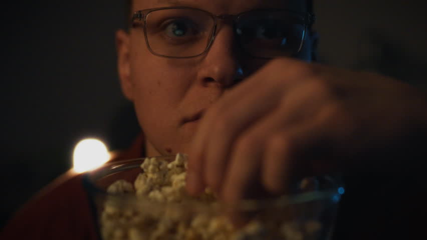 A person sits in a dark space, eyes wide with suspense while reaching for popcorn. The atmosphere is tense as they become engrossed in the movie playing, highlighting their engagement and excitement