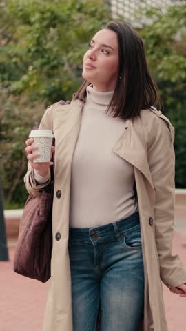 Vertical video, Young woman walking in the city park wearing a trench coat and holding a coffee cup. She's enjoying her peaceful morning walk.