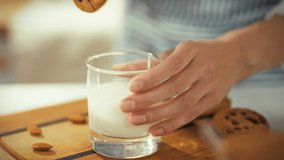 A person is dipping a chocolate chip cookie into a glass of milk on a wooden surface. The warm kitchen setting suggests a cozy snack time, perfect for enjoying a treat - Powered by Shutterstock - Get 15% off with code: PIKWIZARD15