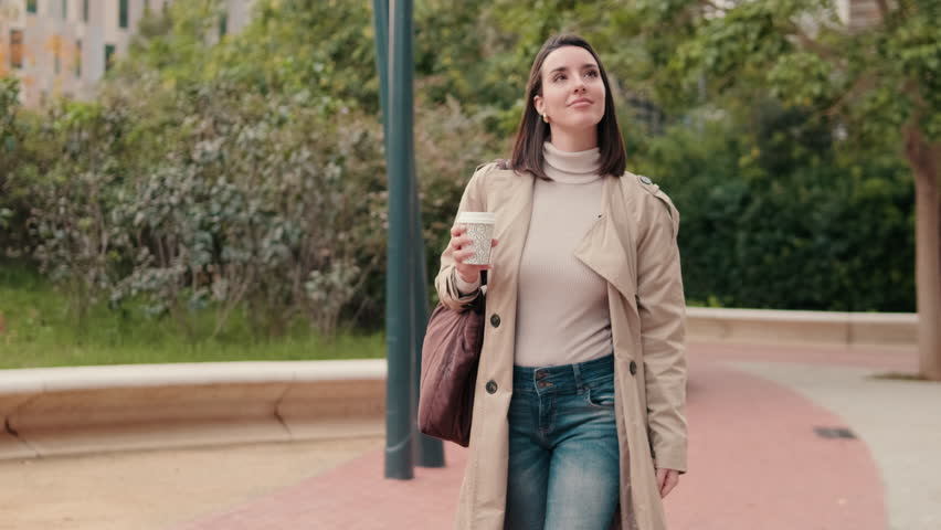 Young woman walking in the city park wearing a trench coat and holding a coffee cup. She's enjoying her peaceful morning walk.