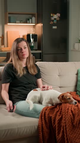 Woman sitting on sofa holding TV remote control. Female watching television at home with pet dog on couch. Concept of relaxation, lifestyle, and domestic life