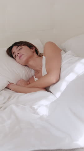 Vertical video, Serene woman sleeps soundly in a sunlit bedroom. White bed linen and soft pillows create a cozy, inviting atmosphere, perfect for relaxation and rest.