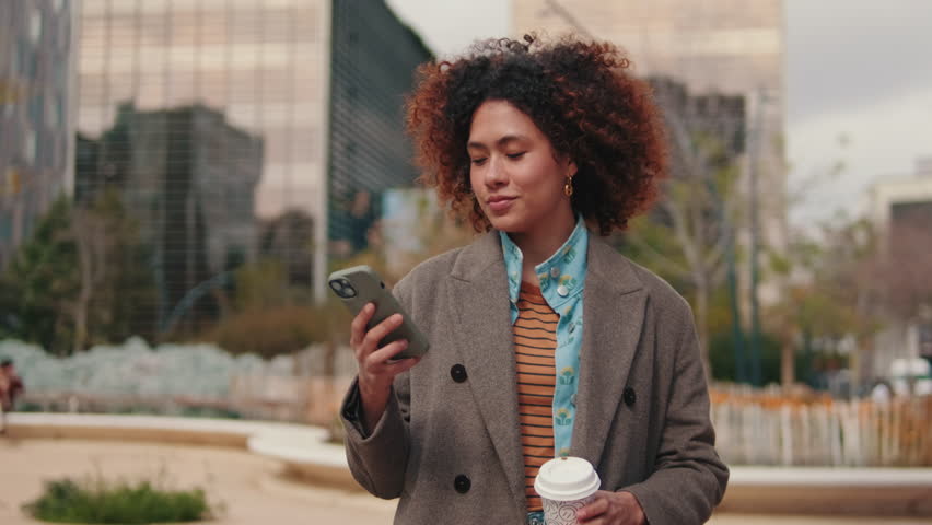 A young woman walks through a city park, enjoying a coffee and browsing her phone. She represents the modern urban lifestyle.