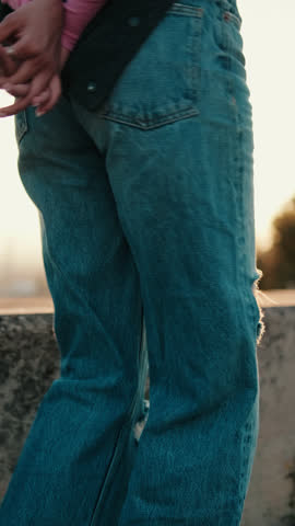 Vertical video, Woman in ripped jeans stands by concrete wall at golden hour. Her backpack rests on the wall as she prepares to dance.
