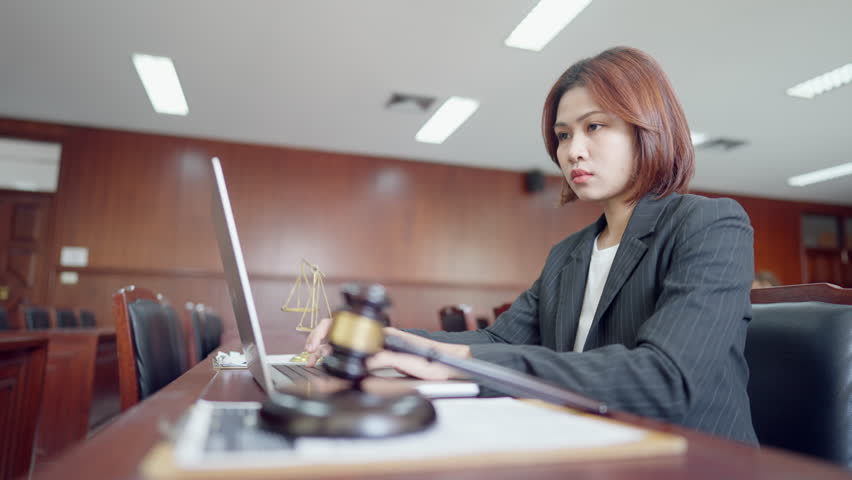 A woman in a suit is sitting at a desk with a laptop and a gavel. She is working on a case or preparing for a trial