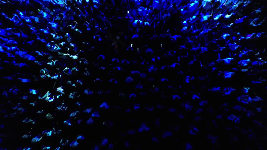 Aerial bird's-eye view of a concert crowd illuminated by stage lights flashing periodically, creating a dynamic atmosphere.