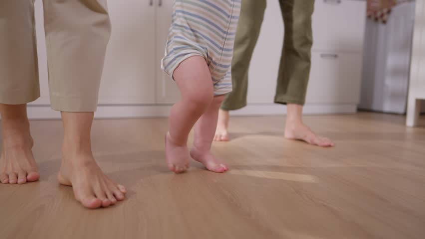 Close up of woman couple cares for and raises their baby boy at home. Two mother share tender moment as they support their baby taking his first steps in kitchen, creating warm family environment.