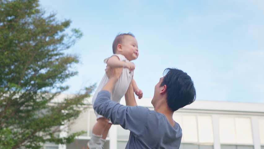 Asian young father holding and gently tosses baby boy outdoors in park. Attractive dad share a tender moment playing with newborn child, fostering a warm and loving family atmosphere in the garden.