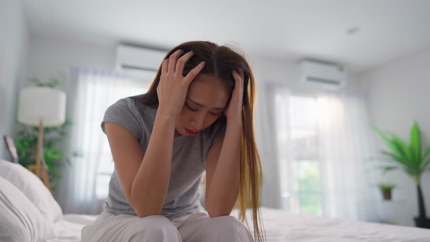 Depressed Asian woman feel upset while sitting alone on bed in bedroom. Attractive frustrated young girl feel sad and headache with life problem and financial in house. Health care Expression Concept.