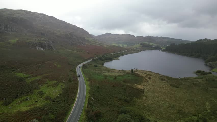 A winding road passes through the rugged Welsh countryside, alongside a tranquil lake and rolling hills. The dramatic, overcast sky adds depth to the scenic landscape, creating a moody view. - Powered by Shutterstock - Get 15% off with code: PIKWIZARD15