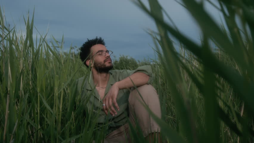 Medium full shot of young polyethnic man in glasses, casual khaki shirt sitting outdoors on cloudy evening among tall green grass with closed eyes, meditating and enjoying feeling of wind on face