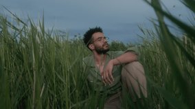 Medium full shot of young polyethnic man in glasses, casual khaki shirt sitting outdoors on cloudy evening among tall green grass with closed eyes, meditating and enjoying feeling of wind on face - Powered by Shutterstock - Get 15% off with code: PIKWIZARD15