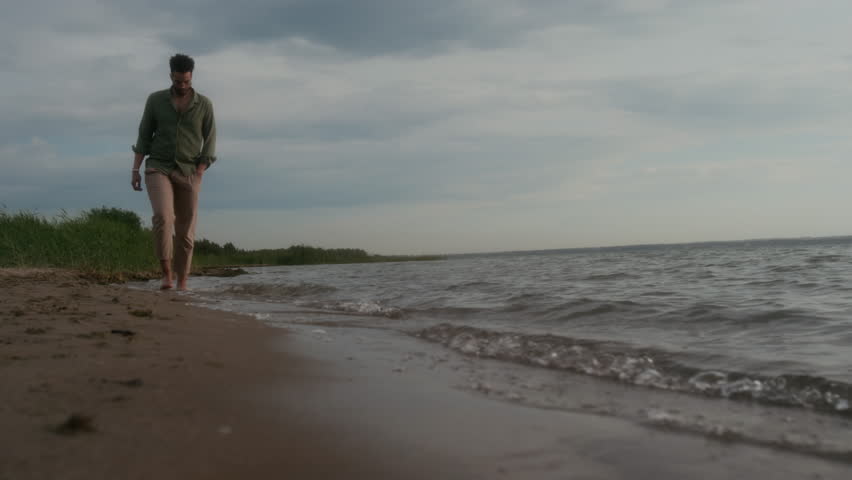 Wide slowmo shot of pensive young African American man with bare feet walking on sandy sea or lake beach, enjoying cool rolling waves, while enjoying time alone outdoors in evening