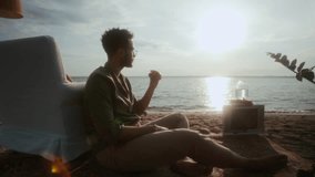 Full shot with lens flare of young man sitting on carpet on sandy sea or ocean beach, eating burger while enjoying sunset picnic in home interior and contemplating solitude - Powered by Shutterstock - Get 15% off with code: PIKWIZARD15