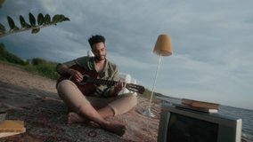 Full rolling shot of solitary young African American man sitting on carpet on sandy beach in evening, with TV, lamp, books, potted plants, playing melancholic melody on guitar - Powered by Shutterstock - Get 15% off with code: PIKWIZARD15
