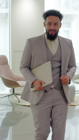 Happy African American businessman in a stylish light suit strides through at modern coworking office with large windows, holding laptop, dancing and having fun after closed amazing deal. 