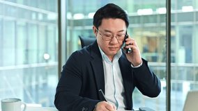 Focused Korean businessman writes notes while talking on phone. Modern office setting with natural light. Professional and efficient worker. - Powered by Shutterstock - Get 15% off with code: PIKWIZARD15