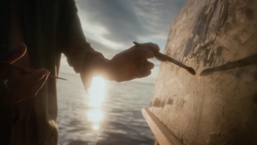 Rolling medium closeup of hand of anonymous male artist holding brush and palette while putting careful finishing touches to oil painting, while working on landscape on sea shore at sunrise