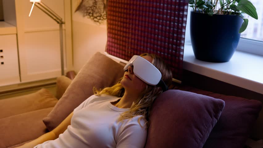 Eye massage device. Young girl wearing glasses for massage relaxing on sofa at home. Woman takes care of her health and skin care