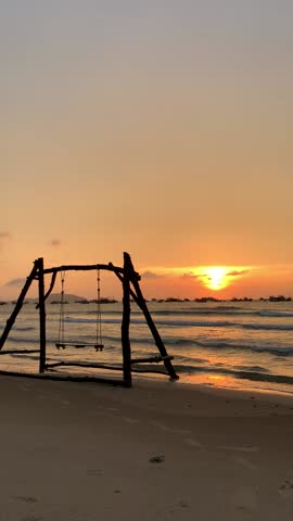 wooden swing hangs between two leaning palm trees on the beach, with the sunset painting the sky in hues of orange and purple, while gentle waves lap at the shore.
