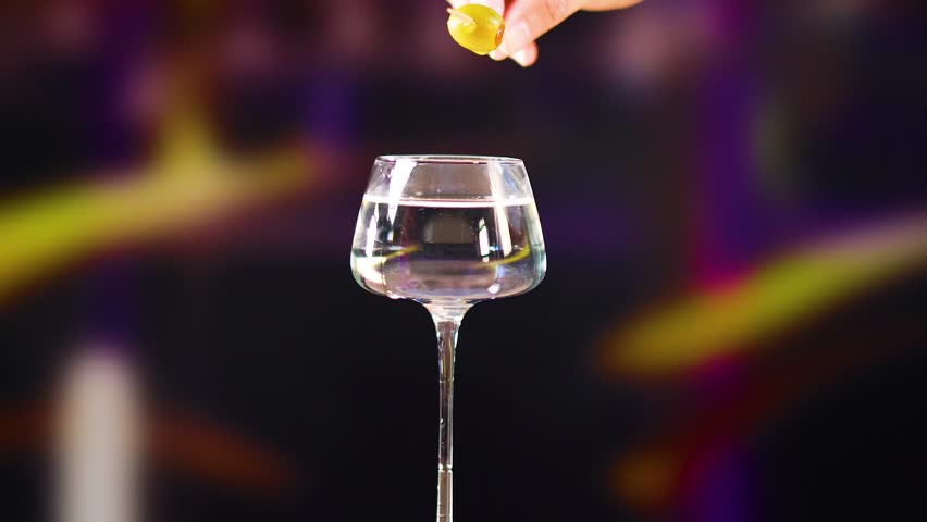 A hand places an olive on a martini glass against a blurred colorful background, highlighting cocktail preparation