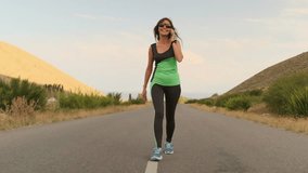 A young woman walks along a road through the hills, answering a call during her workout. Holding the phone to her ear, she continues forward as the peaceful evening landscape stretches around her. - Powered by Shutterstock - Get 15% off with code: PIKWIZARD15