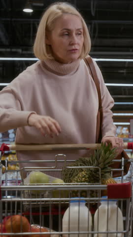 Vertical shot of blonde adult woman choosing fresh bread in bakery at grocery store