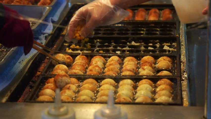 Close up shot of a street vendor cooking takoyaki balls on hot molded griddle, adding ingredients, flipping side making sure every octopus balls are cooked evenly at night market.