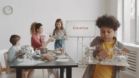 Medium shot of curly biracial boy standing in foreground of teacher and students in classroom, presenting his crafted school project and talking on camera - Powered by Shutterstock - Get 15% off with code: PIKWIZARD15