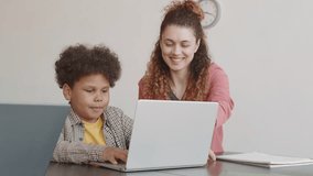 Tilting up slow motion of young Caucasian female teacher helping elementary-aged biracial boy sitting at table, using computer on lesson, smiling - Powered by Shutterstock - Get 15% off with code: PIKWIZARD15