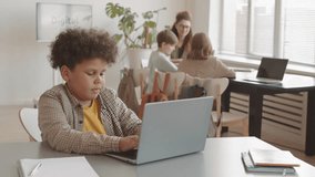Chest-up of cute biracial boy sitting at desk, typing on portable computer in foreground of teacher talking to students in classroom - Powered by Shutterstock - Get 15% off with code: PIKWIZARD15