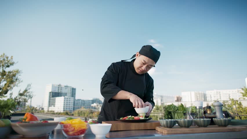 Asian chef adding ingredients to plate and seasoning outdoor. Man in uniform garnishing salad on rooftop. Fresh vegetables and spices on table. Urban background, creating culinary atmosphere.