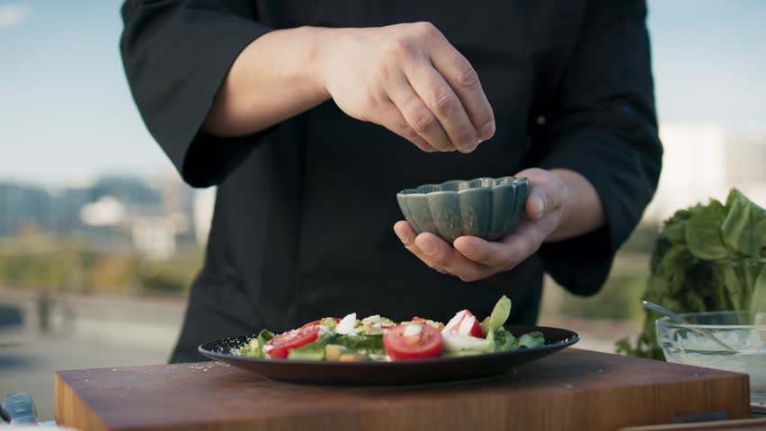 Close-up of Asian chef sprinkling salt over salad on plate outdoors. Rooftop cooking with fresh vegetables, vibrant colors. Professional hands seasoning food. Culinary preparation in open-air kitchen.