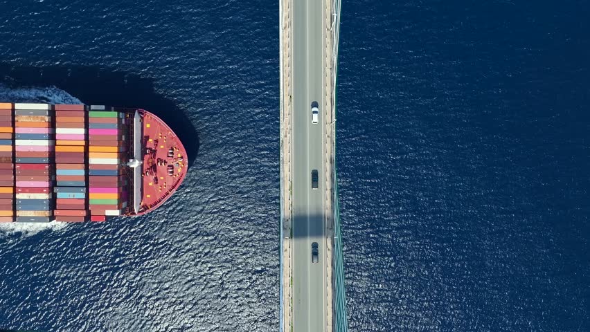 Transport concept with an aerial overhead view of a very big container super cargo ship passing under a bridge with car traffic