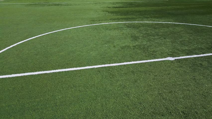 Low-altitude drone shot of an empty soccer field with artificial green grass, white boundary lines, and a goal net. The sports ground is well-maintained, creating a clean and structured composition
