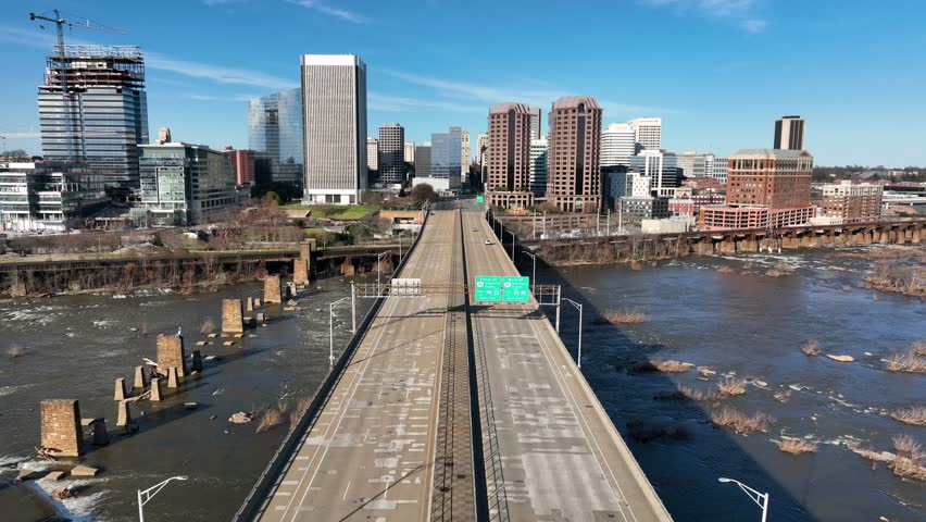 Vehicles on bridge leading to downtown of Richmond, VA. Aerial backwards wide shot. Sunny day in spring. White car crossing James River. Skyline and skyscrapers in distance. Virginia, USA