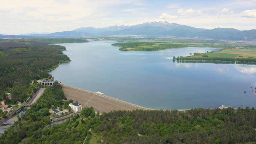 Pedestal drone shot tilting downwards while showing the mountains, lake, and the Koprinka Dam in Central Bulgaria.