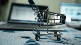 Miniature Shopping Cart on Desk with Financial Charts, Miniature shopping cart on a desk with financial charts and a laptop in the background - Powered by Shutterstock - Get 15% off with code: PIKWIZARD15