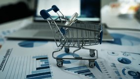 Miniature Shopping Cart on Desk with Financial Charts, Miniature shopping cart on a desk with financial charts and a laptop in the background - Powered by Shutterstock - Get 15% off with code: PIKWIZARD15