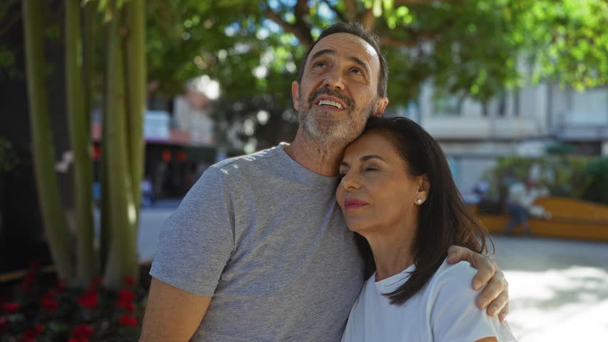 Middle-aged hispanic couple embracing lovingly outdoors in a sunny urban park setting, capturing warmth, happiness, and companionship in an idyllic city environment.