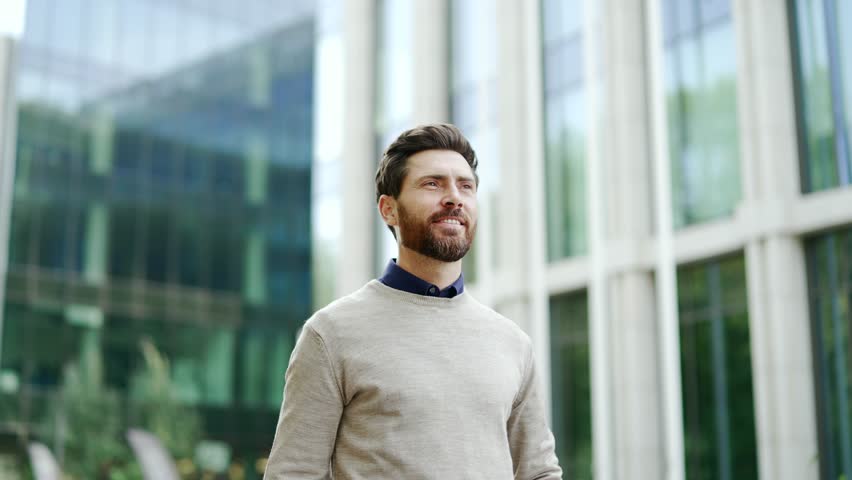 Business professional confidently walks outdoors in cityscape near office building. Mature, bearded man enjoys modern urban environment, exuding positivity, success. business life in corporate setting